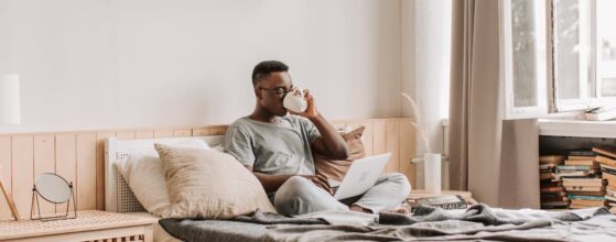 A man working on a laptop and drinking coffee on a bed in a fully furnished temporary housing apartment.